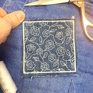 May include: A blue denim patch with a white embroidered floral design. The patch is pinned to a larger piece of blue denim.
