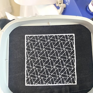 May include: A black fabric square with a white geometric design is held in an embroidery hoop. The design features a repeating pattern of intersecting lines, creating a modern, abstract look. Embroidery machine parts are visible in the background.
