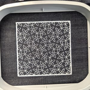 May include: A square of dark denim fabric is held in an embroidery hoop. The fabric features a white floral pattern stitched in a repeating design. The embroidery is framed by a white border.