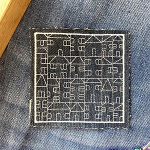 Könnte beinhalten: Ein quadratischer, gestickter Aufnäher mit einer weißen Strichzeichnung von Häusern auf dunkelblauem Hintergrund, auf Denim genäht. Ein Holzobjekt und eine dekorative Schere befinden sich im Hintergrund. Misst ca. 5 cm x 5 cm.