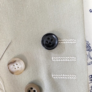 Puede incluir: Primer plano de una tela verde claro con un botón negro cosido. Se ven otros dos botones, uno marrón claro y otro blanquecino. También hay hilo blanco y costuras decorativas.