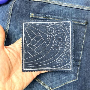 Puede incluir: Un parche cuadrado azul marino con un diseño bordado en blanco de montañas y olas oceánicas. El parche tiene un borde blanco y un borde decorativo festoneado. El parche se muestra frente a unos vaqueros azules.