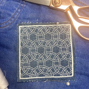 May include: A close-up of a denim patch with a white geometric pattern sewn onto a blue denim fabric. A pair of gold scissors and a spool of white thread are visible in the background.