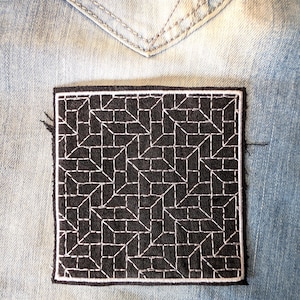 May include: A square, black embroidered patch with a white geometric design, stitched onto a light blue denim fabric. The patch has a white border and is placed near a denim pocket.