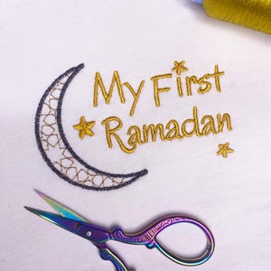 May include: A white t-shirt with gold embroidery of a crescent moon and stars, and the text "My First Ramadan". A pair of rainbow-colored scissors and a spool of gold thread are also visible.