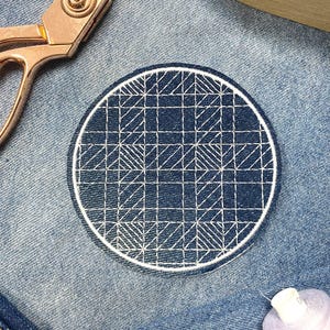 Könnte beinhalten: Ein runder, blauer Jeans-Patch mit einem weißen geometrischen Muster. Der Patch ist mit weißem Garn gestickt und hat einen weißen Rand.