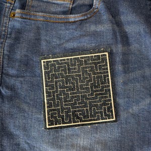 Pode incluir: Um remendo bordado preto e branco com um design de labirinto em um fundo de jeans azul.