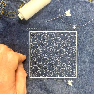 May include: A close-up of a denim fabric with a white embroidered design of swirling lines. The design is framed with a white border. A white spool of thread and a needle with a white butterfly are visible in the background.