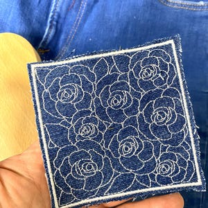 May include: Square denim patch with a white floral rose pattern. The patch has a white border and frayed edges. The patch is held in a hand, with blue jeans in the background.