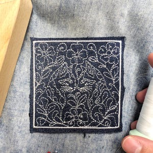 May include: A square dark blue denim patch featuring white embroidered line art of two cats surrounded by floral and leafy patterns. The patch is stitched onto light blue denim fabric, with a spool of white thread and two sewing pins nearby.