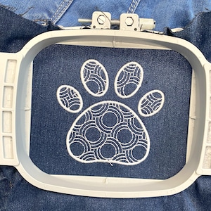 May include: A white paw print embroidery design with a circular pattern inside each toe, stitched onto a dark blue denim fabric in an embroidery hoop.
