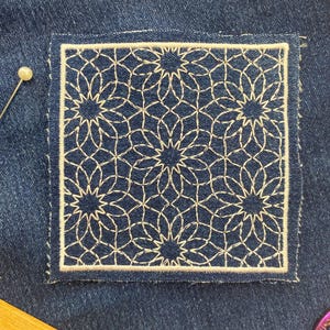 Puede incluir: Parche cuadrado de mezclilla con un diseño floral bordado en blanco. El diseño presenta un motivo repetitivo en forma de estrella dentro de un borde blanco. Un alfiler con cabeza de perla se coloca cerca del parche, lo que sugiere un proyecto de costura.