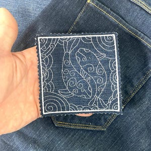 May include: Square denim patch with a white embroidered design of two stylized fish and swirling patterns. The patch has a white border and is held over a pair of blue jeans.