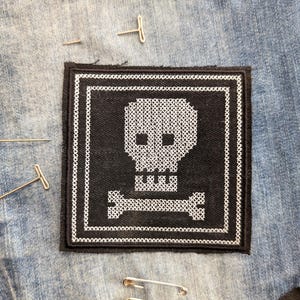 May include: A black and white cross-stitch patch featuring a skull and crossbones design. The patch is square with a white border and is placed on a denim background. Several pins and safety pins are scattered around the patch.