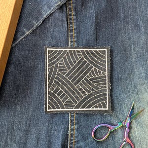 Könnte beinhalten: Ein quadratischer, schwarz-weißer, gestickter Aufnäher mit geometrischem Design ist auf eine Jeansjacke genäht. Eine Regenbogenfarbene Schere liegt daneben. Ein Holzobjekt ist in der oberen linken Ecke sichtbar.