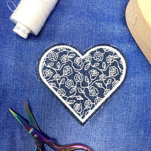 Può includere: Una toppa a forma di cuore bianca con un motivo floreale ricamato in filo nero su uno sfondo di denim blu. Sono visibili anche un rocchetto di filo bianco e un paio di forbici iridescenti.