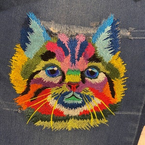 May include: A colorful embroidered cat face with yellow, green, blue, red, and pink thread. The cat's eyes are blue and the nose is red. The cat's whiskers are yellow.