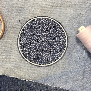 Puede incluir: Un parche circular bordado en azul marino con un diseño blanco en espiral, cosido sobre tela vaquera. El parche tiene un borde blanco. Se ven suministros de costura, incluyendo hilo y tijeras.