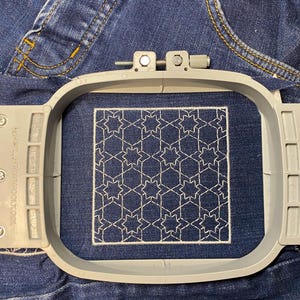 Op de afbeelding: Een close-up van een wit geometrisch patroon geborduurd op een stuk blauwe denim stof. De stof is strak gespannen in een ronde borduurring.