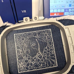 Puede incluir: Una tela de mezclilla azul oscuro sujeta en un bastidor de bordado gris claro, con un contorno bordado en blanco de un perro rodeado de varios patrones florales. El bordado se encuentra bajo la pantalla de una máquina de bordar azul y blanca.