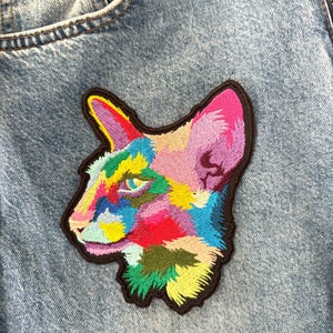 Op de afbeelding: Geborduurde patch met een kleurrijk kattenkop-profiel. De patch toont een levendig kleurenpalet met roze, geel, blauw en groen, omlijnd met een bruine rand. De patch is bevestigd op een denim kledingstuk.