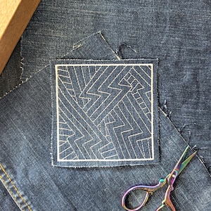 Könnte beinhalten: Ein quadratischer Denim-Patch mit einem weißen geometrischen Design, das mit weißem Faden genäht wurde. Der Patch befindet sich auf einem Denim-Hintergrund, mit einer schillernden Schere. Das Design zeigt eine Reihe von Zickzacklinien und geraden Linien.