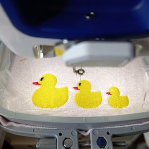 May include: A close-up of a white embroidery machine with a hoop containing three yellow embroidered rubber duckies. The duckies are different sizes, with the largest one on the left and the smallest one on the right.