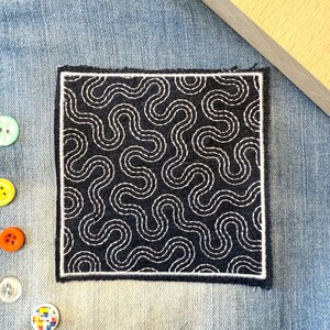 May include: A square, dark blue patch with a white, wavy, embroidered design. The patch is bordered with a white stitched outline. Various colorful buttons and a safety pin are scattered around the patch on a denim background.