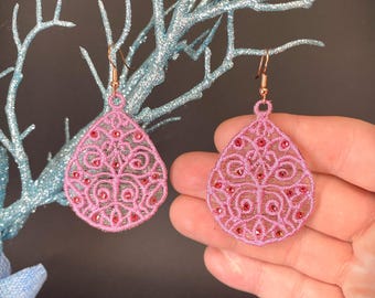 May include: A pair of pink dangle earrings with a delicate, intricate design. The earrings are made of a lightweight material and feature a small, clear crystal in the center of each design.