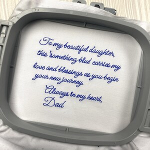 Peut inclure: Tissu blanc brodé avec du fil bleu, portant un message touchant. Le texte dit : « À ma belle fille, ce quelque chose de bleu porte mon amour et mes bénédictions alors que tu commences ton nouveau voyage. Toujours dans mon cœur, Papa. »