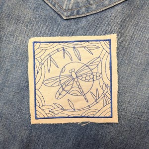May include: A square fabric patch featuring blue machine embroidery of a dragonfly centered within a circular frame, surrounded by bamboo stalks and curved lines, all stitched onto a blue denim fabric background with a visible pocket seam.