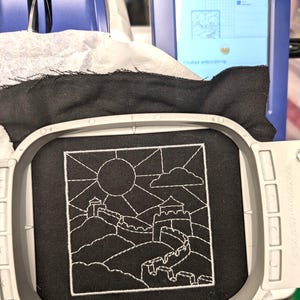 May include: A square embroidery hoop holds black fabric featuring a white line-art design of the Great Wall of China. The scene includes a sun, a cloud, and a watchtower on a hilly landscape. The embroidery machine screen displays 97.0mm by 97.0mm.
