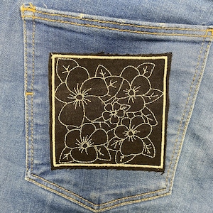Op de afbeelding: Close-up van een blauwe spijkerbroek met een achterzak. Een zwart vierkant patch met een wit bloemenontwerp is op de zak genaaid. De patch heeft een cluster van bloemen en bladeren.