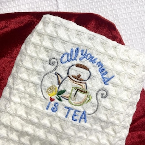Puede incluir: Toalla de cocina blanca con tejido de gofre con un diseño bordado con temática de té. El diseño incluye una tetera, una taza de té, una rodaja de limón y el texto "All you need is TEA" en hilo azul y verde. La toalla está sobre una superficie roja.