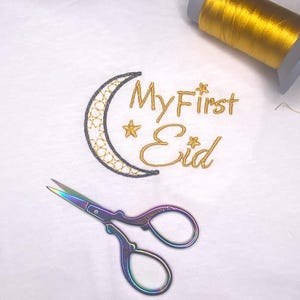 Può includere: Tessuto bianco con ricamo dorato "My First Eid" e un design a mezzaluna. Sono visibili anche un paio di forbici iridescenti e una bobina di filo dorato.