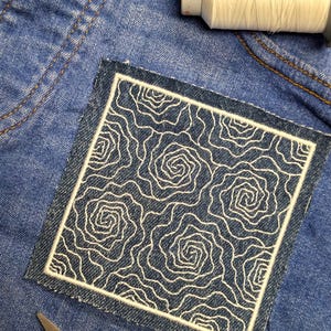 May include: A square denim patch with a white embroidered rose pattern. The patch is bordered with white stitching and is set on a blue denim background. A spool of white thread and scissors are also visible.