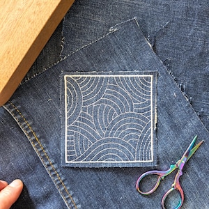 Könnte beinhalten: Ein quadratischer Denim-Patch mit einem weißen, gestickten Wellenmuster auf einem Denim-Hintergrund. Ein Holzblock und eine Regenbogen-Schere sind ebenfalls vorhanden. Der Patch dient wahrscheinlich zur Reparatur oder Individualisierung von Kleidung.
