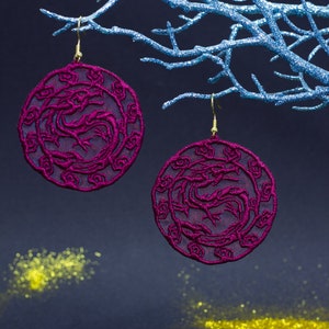 May include: A pair of dangle earrings with a circular design featuring a dragon in a pink color. The earrings are hanging from a blue branch.