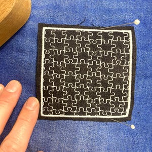 Pode incluir: Um remendo de tecido preto com um desenho de quebra-cabeça branco. O remendo está preso a um tecido de ganga azul.