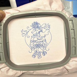 May include: An embroidery hoop holding pink fabric with a blue embroidered design. The design includes a jar with the words "Just Married", doves, and floral elements. The embroidery is in progress, showing the stitching.