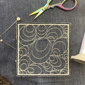 May include: A denim patch featuring a white embroidered swirl pattern. The patch is set on a denim background, with a pair of rainbow scissors, two pins, and a white thread spool. Suitable for sewing or craft projects.