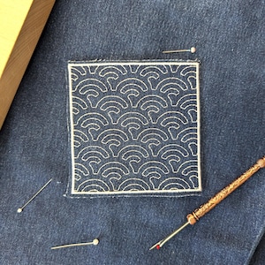 Może przedstawiać: Zbliżenie na dżinsową tkaninę z kwadratową naszywką z białym wzorem fal. Kilka szpilek i szewc rozsiane wokół naszywki. Drewniane narzędzie widoczne w lewym górnym rogu.