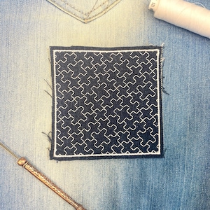 May include: A square, dark blue patch with a white geometric pattern is sewn onto a light blue denim surface. A white thread spool and a gold-colored sewing tool are also visible.
