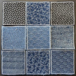 Puede incluir: Nueve parches cuadrados de tela con diseños bordados. Los parches presentan varios patrones, incluyendo formas geométricas, espirales y motivos de olas. El bordado es blanco sobre un fondo de mezclilla azul oscuro, con un borde blanco alrededor de cada cuadrado.