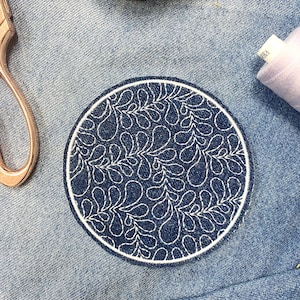 May include: A round, navy blue embroidered patch with a white border and a repeating white leaf pattern. The patch is on a denim background, with a pair of scissors and a wooden object in the background.