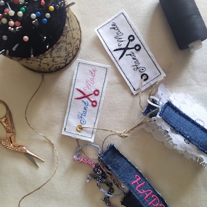 Puede incluir: Primer plano de una pulsera de mezclilla con bordado rosa que dice "Happy". La pulsera está atada con un cordel a dos etiquetas de tela blanca con bordado negro que dice "Hand Made" y un par de tijeras. También se puede ver un carrete de hilo negro y un alfiletero con alfileres de colores.