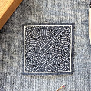 Könnte beinhalten: Ein quadratischer, marineblauer Stoffflicken mit einem weißen, wirbelnden Stickmuster. Der Flicken befindet sich auf einem Jeanshintergrund, mit einem Holzblock und einem Nahttrenner im Bild.