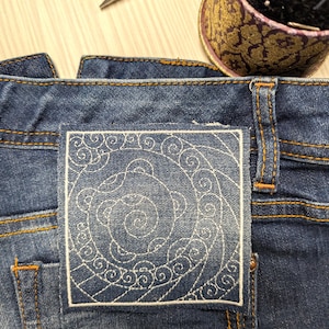 Peut inclure: Un écusson carré en jean avec des motifs brodés blancs en forme de spirales et de tourbillons, cousu sur le dos d'un jean bleu foncé avec des surpiqûres orange. Une paire de ciseaux en métal et un pelote à épingles sont visibles en haut.