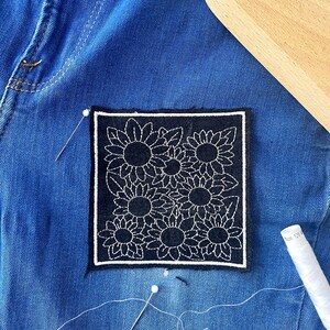 May include: A square, black patch with a white sunflower design is sewn onto blue denim. White thread, needles, and a wooden tool are also visible. The patch is embroidered with a detailed floral pattern.