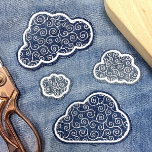 May include: Four white embroidered patches with a blue swirl pattern in the shape of clouds. The patches are on a blue denim background.
